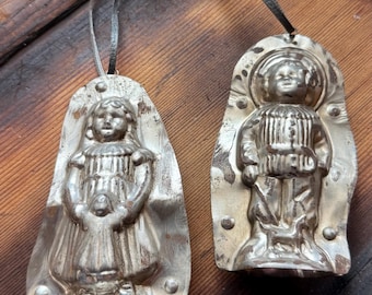 May include: Two vintage silver-toned metal molds shaped like figures, each featuring a hanging loop at the top. One mold depicts a person in a dress, and the other depicts a person in a collared jacket and trousers, both with embossed details.