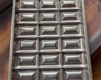 May include: A rectangular silver-colored metal chocolate mold featuring a grid of twenty-four individual recessed rectangular compartments arranged in four columns and six rows, designed for creating uniform chocolate bar segments.