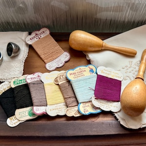 May include: A collection of vintage sewing supplies, including spools of thread in various colors, wooden tools, a pin cushion, and a thimble. The threads are labeled with the words "EMBROIDERY" and "PURE DYE". The items are arranged on a wooden surface.