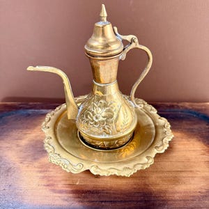 May include: A polished brass oil lamp with a long, slender spout, a curved handle, and a pointed lid. The body of the lamp features intricate floral engravings. It rests on a matching round brass tray with a decorative, scalloped, embossed edge.