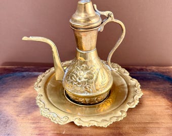 May include: A polished brass oil lamp with a long, slender spout, a curved handle, and a pointed lid. The body of the lamp features intricate floral engravings. It rests on a matching round brass tray with a decorative, scalloped, embossed edge.