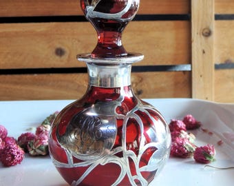 May include: A ruby red glass perfume bottle with a silver overlay design. The bottle has a round body and a bulbous stopper. The silver overlay features floral and abstract patterns. The bottle is displayed on a white tray with dried flowers.