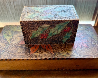 May include: Two wooden boxes, one stacked on the other. The top box is square and features a holly leaf and berry design. The bottom box is rectangular with a leaf design. Both boxes have a dark brown background with colorful accents.