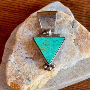 May include: A silver pendant with a triangular turquoise stone. The turquoise is a vibrant teal color with a cracked pattern. The pendant has a rectangular bail and three small silver spheres at the bottom.