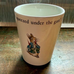 May include: Cream-colored ceramic mug featuring a cartoon illustration of a rabbit in a blue jacket holding carrots. The mug has the text "squeezed under the gate!" printed in black.