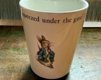 May include: Cream-colored ceramic mug featuring a cartoon illustration of a rabbit in a blue jacket holding carrots. The mug has the text "squeezed under the gate!" printed in black.