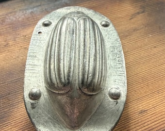 May include: A silver-toned metal mold featuring a raised, elongated beetle or insect shape with prominent vertical ridges on its back. The mold has an oval-shaped flat rim with four small circular indentations at the corners and a textured surface.