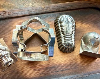 May include: A collection of four metal kitchen molds and cutters. The set includes a small Santa Claus mold, a large geometric cookie cutter, a textured lobster-shaped mold, and a rounded dome-shaped mold, all crafted from silver-toned metal.