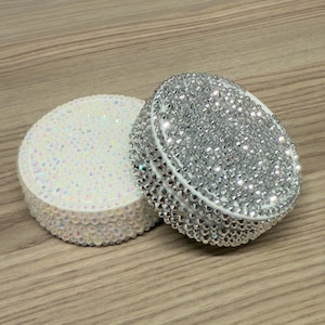 May include: Two round, bejeweled containers. One is white with iridescent gems, the other is silver with clear crystals. Both have a white rim and are set on a light brown wooden surface. The containers are likely for storing small items.
