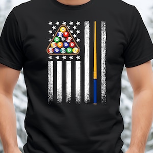 May include: Black t-shirt featuring a graphic of the American flag with a pool table theme. The flag design incorporates a triangle of billiard balls in place of the stars and a pool cue in place of the stripes. The balls are numbered.