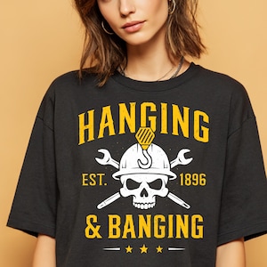 Puede incluir: Camiseta negra con un gráfico amarillo y blanco. El gráfico incluye las palabras "HANGING & BANGING" sobre una calavera con un casco y llaves cruzadas. También se incluye el texto "EST. 1896".