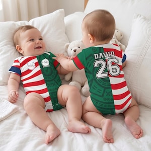 Puede incluir: Dos bebés con mamelucos a juego rojos, blancos, azules y verdes con un logotipo. Un mameluco tiene el nombre "DAVID" y el número "26" en la espalda. Los bebés están acostados en una cama blanca con almohadas y un osito de peluche.