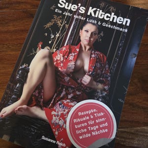 Könnte beinhalten: Buchcover von "Sue's Kitchen" mit einer Frau in einem roten Blumenmuster-Kimono. Der Titel ist in weißen Buchstaben, mit deutschem Text. Ein Aufkleber wirbt für Rezepte und Rituale.
