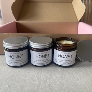 May include: Three glass jars of honey-scented candles with silver lids, labelled "The Selection Honey." The jars are in a cardboard box with a pink interior. Each candle is designed for a 22-hour clean burn.