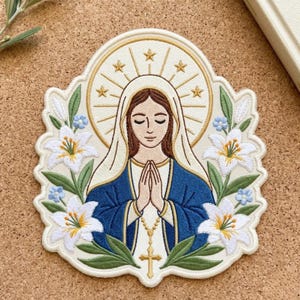 May include: An embroidered patch featuring a figure with brown hair wearing a white veil and blue robe, hands clasped in prayer with a gold rosary. The figure is surrounded by white lilies, blue flowers, and a gold halo with stars.