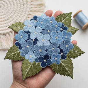 May include: An embroidered patch featuring a cluster of hydrangea flowers in various shades of blue, ranging from light sky blue to deep navy, accented with detailed green leaves with visible stitching and serrated edges.