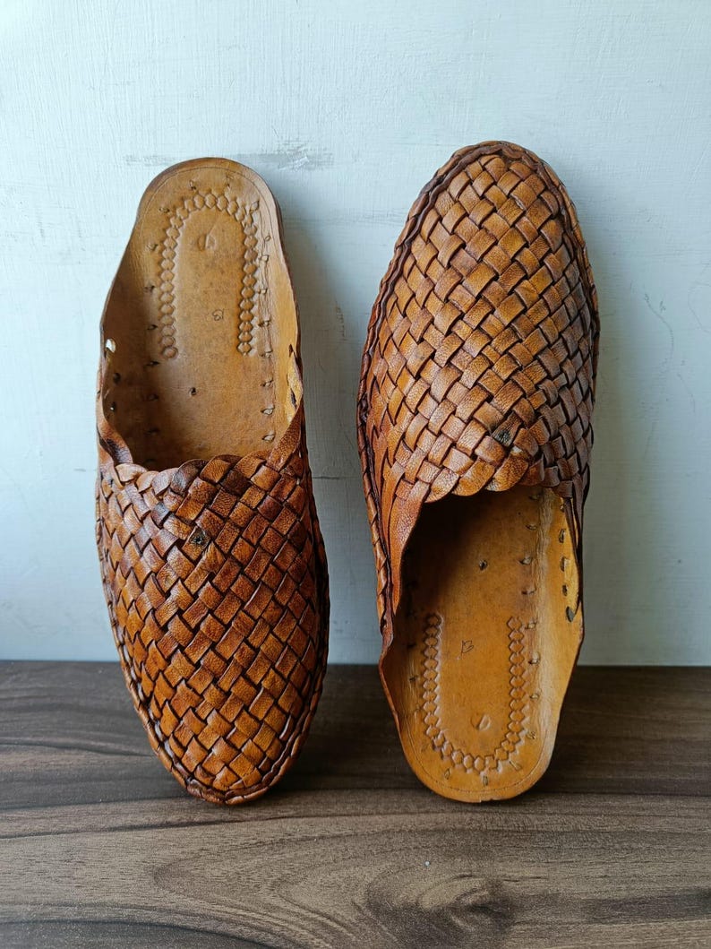 May include: A pair of brown leather slip-on shoes. The shoes feature a woven design on the top and a leather sole. The shoes are displayed on a wooden surface against a white background. The shoes are a traditional style.