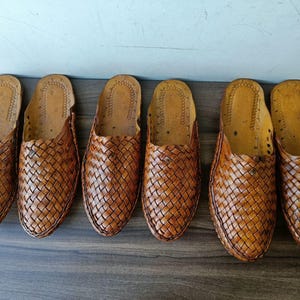 May include: Six pairs of brown leather woven slip-on shoes are arranged in a row. The shoes have a textured, basket-weave design on the upper and a smooth, light-colored interior. The shoes are displayed on a dark wooden surface.