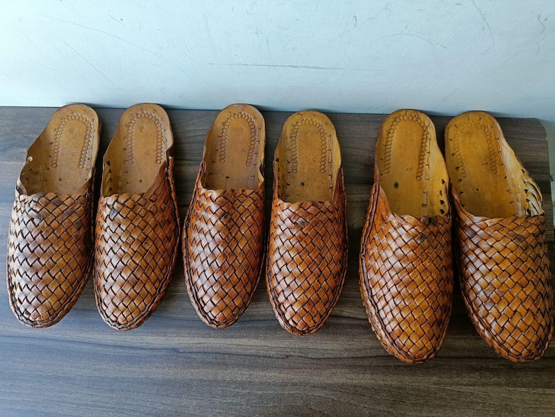 May include: Six pairs of brown leather woven slip-on shoes are arranged in a row. The shoes have a textured, basket-weave design on the upper and a smooth, light-colored interior. The shoes are displayed on a dark wooden surface.