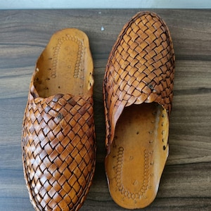 May include: A pair of brown leather slip-on shoes. The shoes feature a woven design on the top and sides. The soles are a lighter shade of brown. The shoes are designed for comfort and casual wear.