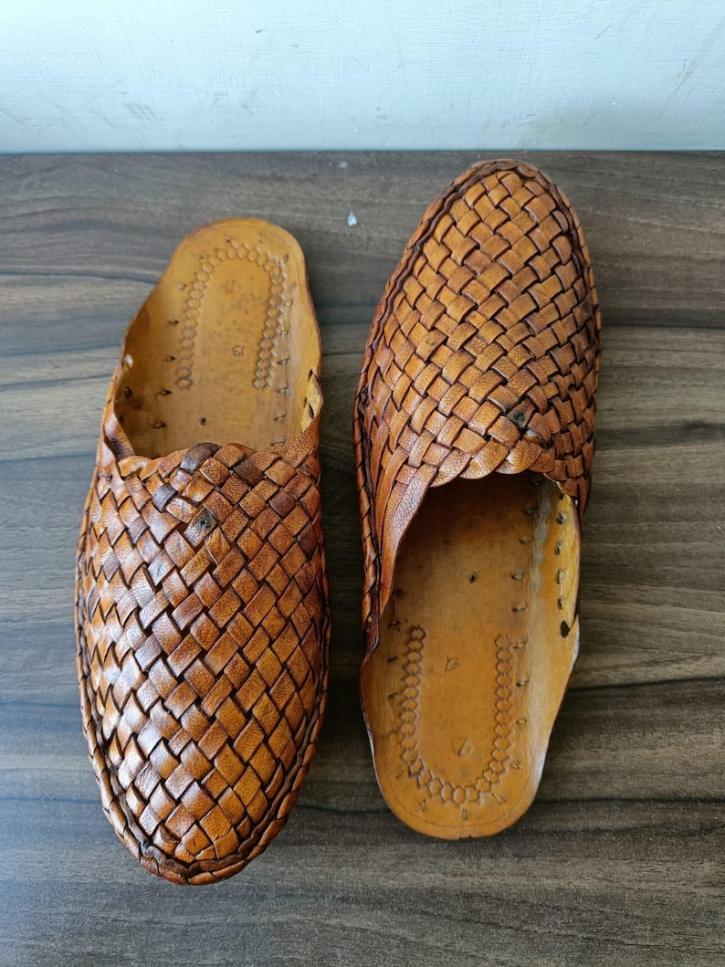 May include: A pair of brown leather slip-on shoes. The shoes feature a woven design on the top and sides. The soles are a lighter shade of brown. The shoes are designed for comfort and casual wear.