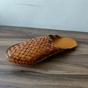 May include: A single brown leather slipper with a woven design. The slipper is made of light brown leather and has a woven pattern across the top. The sole is a darker shade of brown. The slipper is designed for indoor use.