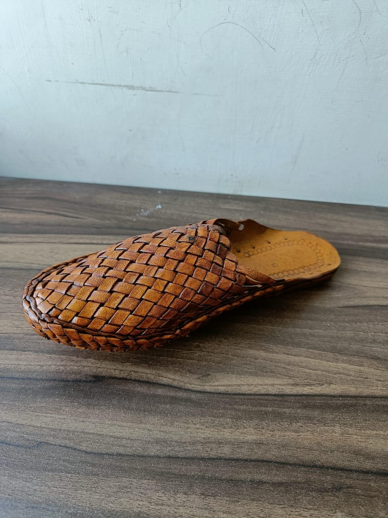 May include: A single brown leather slipper with a woven design. The slipper is made of light brown leather and has a woven pattern across the top. The sole is a darker shade of brown. The slipper is designed for indoor use.