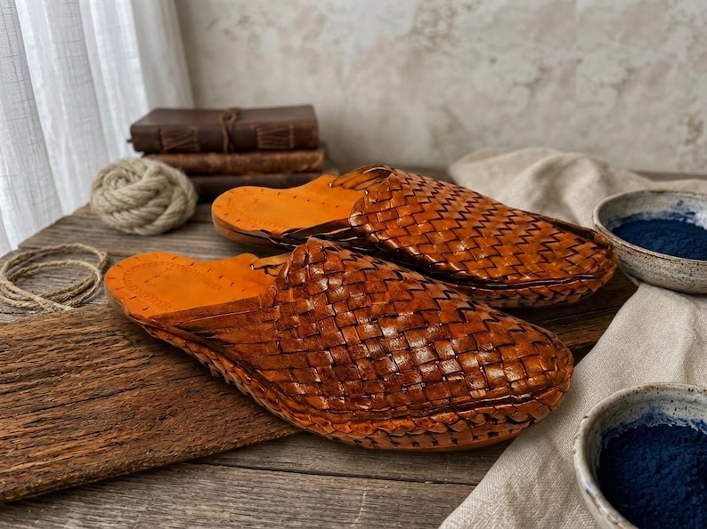 May include: A pair of tan brown leather mules featuring a woven basket-weave pattern across the upper section. The interior footbed is a smooth, lighter tan leather with visible stitching along the edges and a rounded toe silhouette.