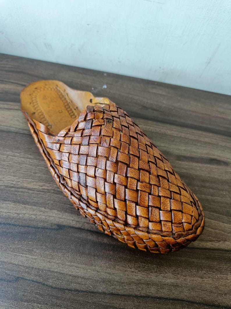 May include: A single brown leather slipper with a woven design. The slipper is made of a light brown leather and has a textured appearance. The slipper is designed for indoor use.
