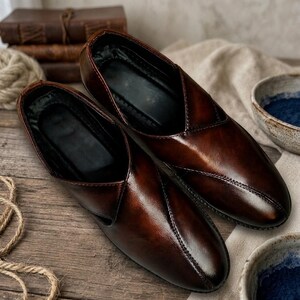 May include: A pair of brown leather slip-on shoes with a glossy finish. The shoes have a pointed toe and a stitched seam detail. The interior is black. The shoes are displayed on a wooden surface.