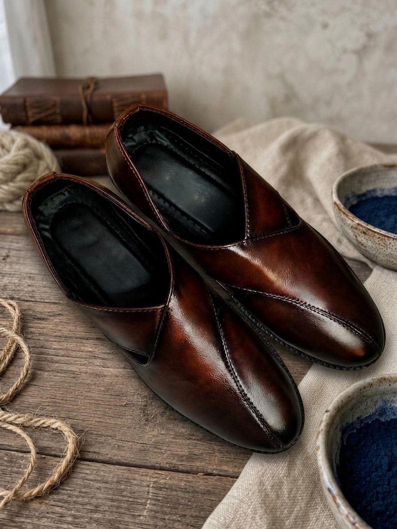 May include: A pair of brown leather slip-on shoes with a glossy finish. The shoes have a pointed toe and a stitched seam detail. The interior is black. The shoes are displayed on a wooden surface.