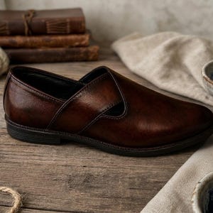May include: A brown leather slip-on shoe with a unique design. The shoe has a dark sole and a smooth, polished finish. The image also shows a stack of books, rope, and bowls.