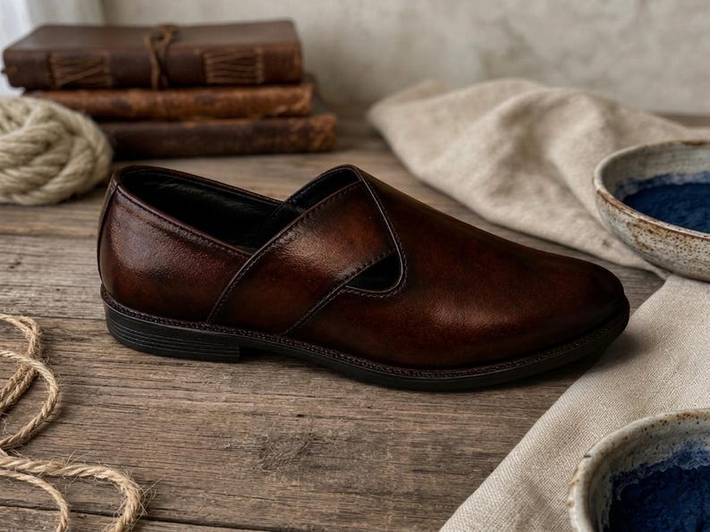 May include: A brown leather slip-on shoe with a unique design. The shoe has a dark sole and a smooth, polished finish. The image also shows a stack of books, rope, and bowls.