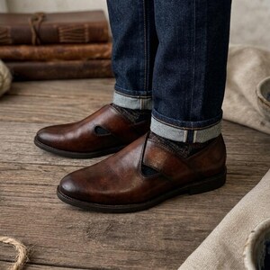 May include: A pair of brown leather slip-on shoes with a cut-out detail on the side. The shoes are worn with dark blue denim jeans with the cuffs rolled up. The background includes wooden planks, books, and rope.
