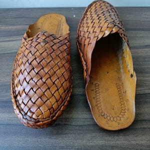 May include: Two brown leather woven slip-on shoes. The shoes have a textured, basket-weave design on the top and a smooth leather sole. The shoes are displayed on a dark wooden surface.