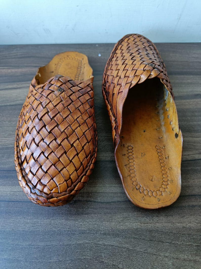 May include: Two brown leather woven slip-on shoes. The shoes have a textured, basket-weave design on the top and a smooth leather sole. The shoes are displayed on a dark wooden surface.