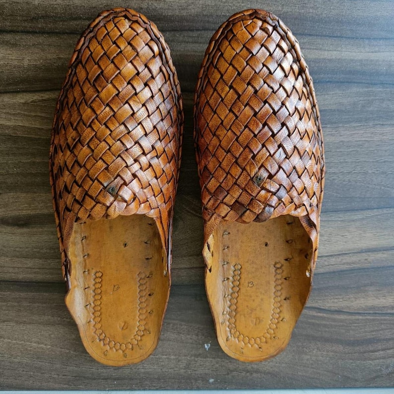 May include: A pair of brown leather woven slip-on shoes. The shoes have a detailed basket weave pattern on the top and a light brown sole. The shoes are displayed on a wooden surface.