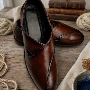 May include: A pair of brown leather shoes with a pointed toe and a low heel. The shoes have a glossy finish and are displayed on a wooden surface. The shoes are a slip-on style and appear to be handmade.
