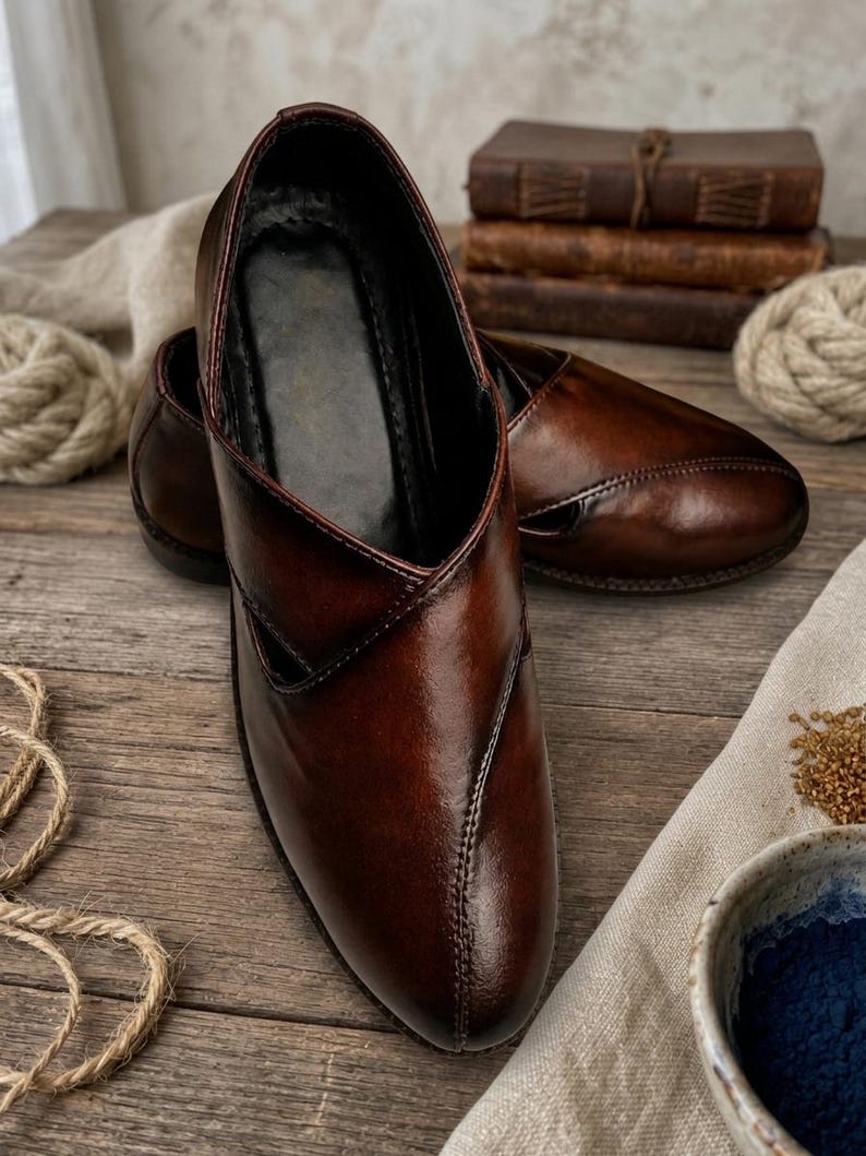 May include: A pair of brown leather shoes with a pointed toe and a low heel. The shoes have a glossy finish and are displayed on a wooden surface. The shoes are a slip-on style and appear to be handmade.