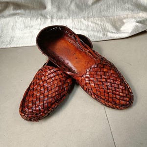 May include: A pair of brown leather slip-on shoes featuring a woven lattice pattern across the upper surface. The shoes have a rounded toe, an open top design, and a smooth, solid leather interior lining with a deep, rich tan color throughout.
