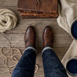 May include: Overhead shot of brown leather boots, dark denim jeans, and a stack of leather-bound journals on a wooden surface. A coil of rope and two bowls of blue powder are also visible, along with a beige cloth.