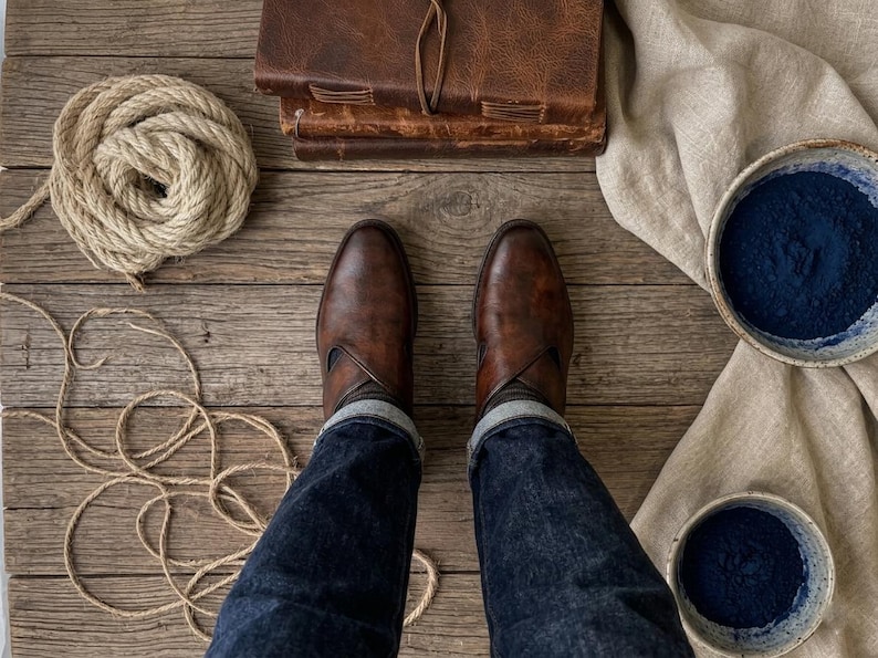 May include: Overhead shot of brown leather boots, dark denim jeans, and a stack of leather-bound journals on a wooden surface. A coil of rope and two bowls of blue powder are also visible, along with a beige cloth.