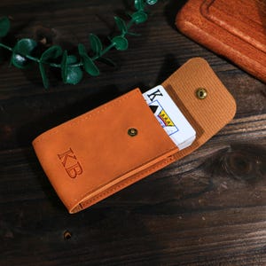 May include: A tan leather card case with the initials "KB" embossed on the front. The case is open, revealing a deck of playing cards with the King of Spades visible. The case is secured with a snap closure.
