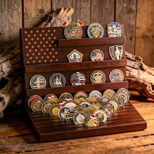 Puede incluir: Un expositor de madera marrón oscuro con múltiples estantes, que muestra una colección de monedas de desafío militares. El expositor presenta un diseño de bandera estadounidense y la inscripción "HM2(FMF/CAC) Linda Miller, USN."
