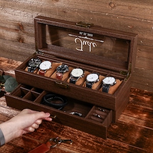 May include: A wooden watch box with a clear glass top, displaying six watches on tan cushions. The box has a drawer below with compartments for accessories. The text "Jude" and numbers are visible on the glass. The box is on a wooden surface.