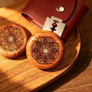 May include: Two circular wooden tools with engraved mandala patterns and a central metal rivet. One tool holds a silver-colored metal razor blade, and both are positioned near a dark red leather pouch with a gold-colored snap button closure.