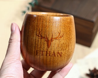 Puede incluir: Una taza de madera con un diseño de cornamenta de ciervo y el nombre "ETHAN" grabado en la parte delantera. La taza es de madera marrón claro y tiene una forma suave y redondeada. La taza se sostiene en una mano.