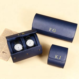 Puede incluir: Estuches y rollos para relojes de cuero azul marino con letras doradas. Un estuche contiene dos relojes con esferas blancas y correas negras. Los otros estuches y rollos tienen las iniciales "FG" y "KJ".