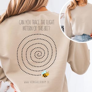 Könnte beinhalten: Beiges Sweatshirt mit einem schwarzen Punkt-Spiralmuster und einer Cartoon-Biene. Der Text "CAN YOU TRACE THE FLIGHT PATTERN OF THIS BEE?" ist über der Spirale aufgedruckt. Die Website-Adresse www.honigheldenvm.de ist darunter gedruckt.