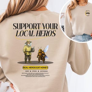Könnte beinhalten: Beiges Sweatshirt mit dem Text "SUPPORT YOUR LOCAL HEROES" und einer Illustration eines Imkers und einer Biene. Unter der Illustration steht der Text "REAL HEROS EAT HONEY". Das Sweatshirt hat auch Text auf Deutsch.